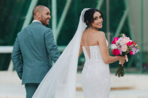 Bride in lace gown holds vibrant bouquet and smiles with groom at Tirtha Uluwatu, a Bali wedding venue.