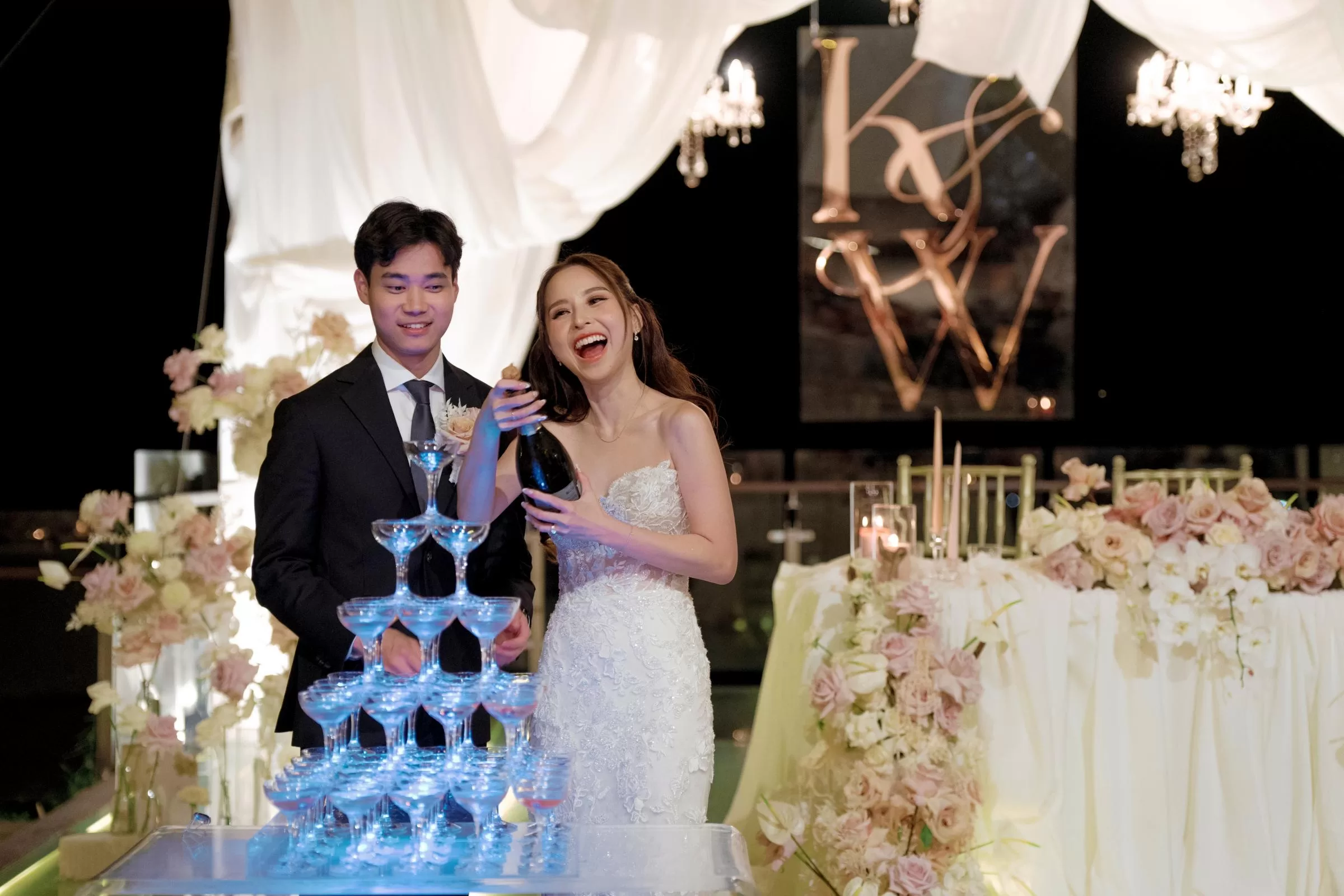 The bride and groom get ready to pour champagne into a stacked tower of glasses at their reception at Tirtha Bali.