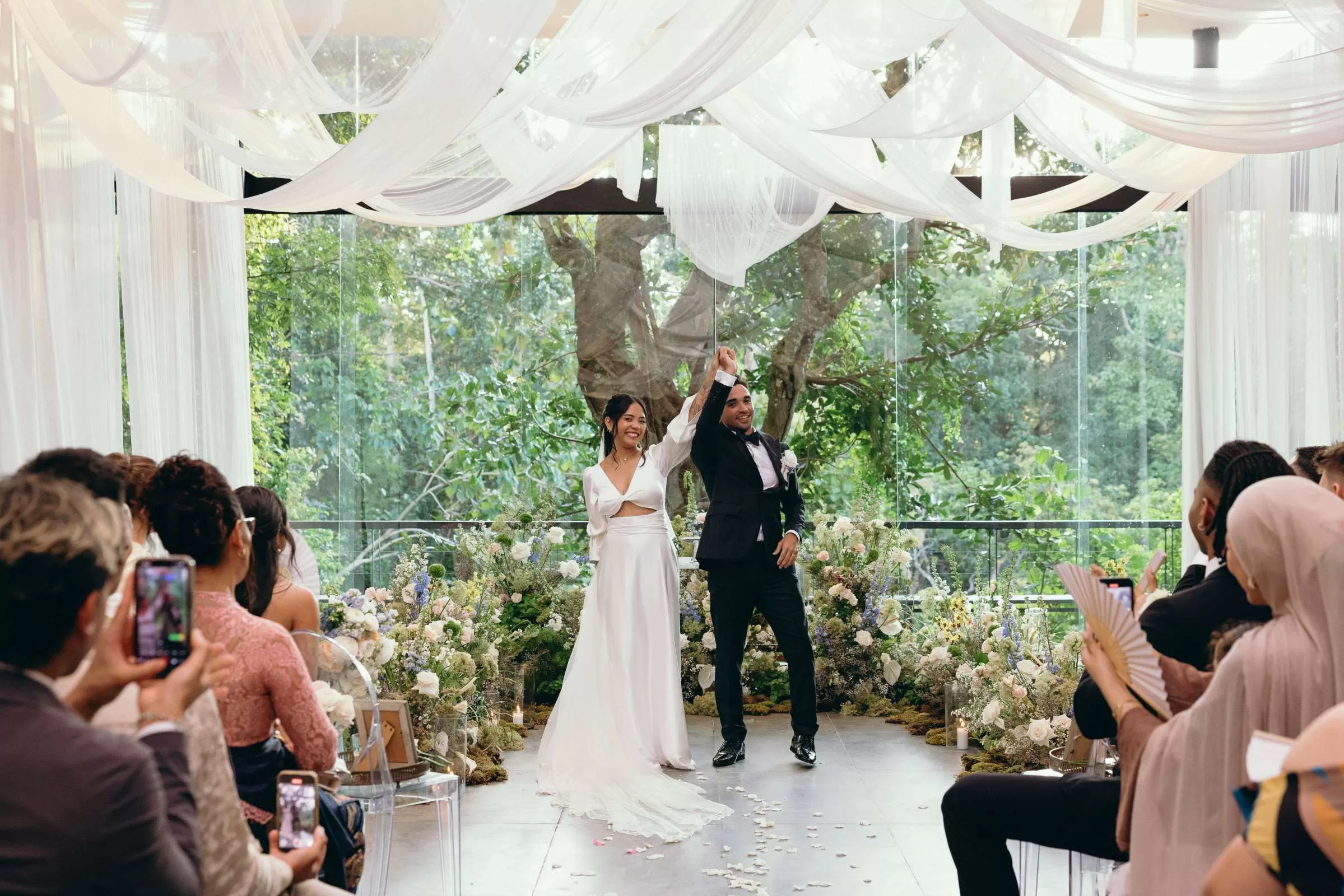 Bride and groom raise their joined hands at the altar, smiling at guests in the Plumeria Room.