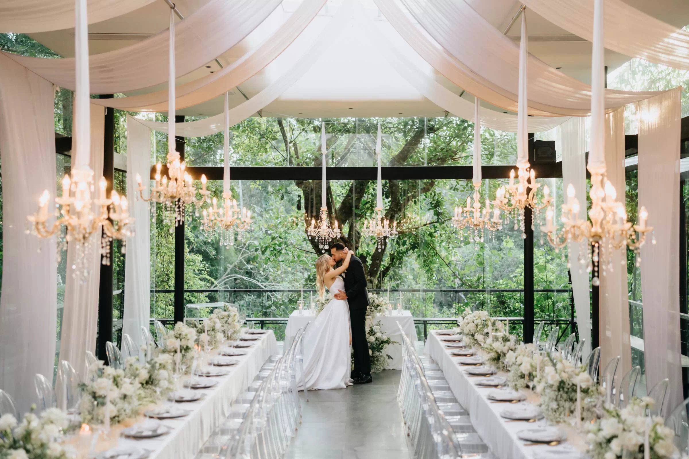 Bride and groom share a kiss in the elegant Plumeria Room, adorned with white decor and chandeliers.