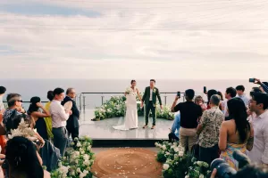 A newlywed couple hold hands on the Glass Stage at Tirtha Uluwatu with an ocean backdrop as the guests cheer and photograph.