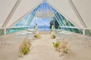 Tirtha Uluwatu’s Glass Chapel as a wedding ceremony venue in the Eve package, with floral aisle decor and ghost chairs.