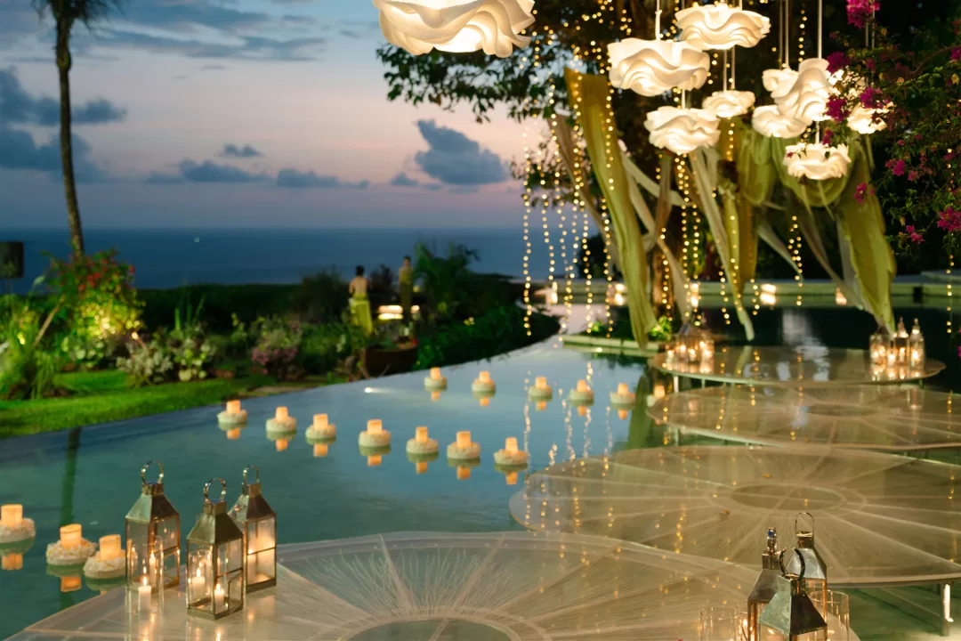 Tirtha Uluwatu's floating stage with floral lamps and candles overlooking the ocean at sunset.