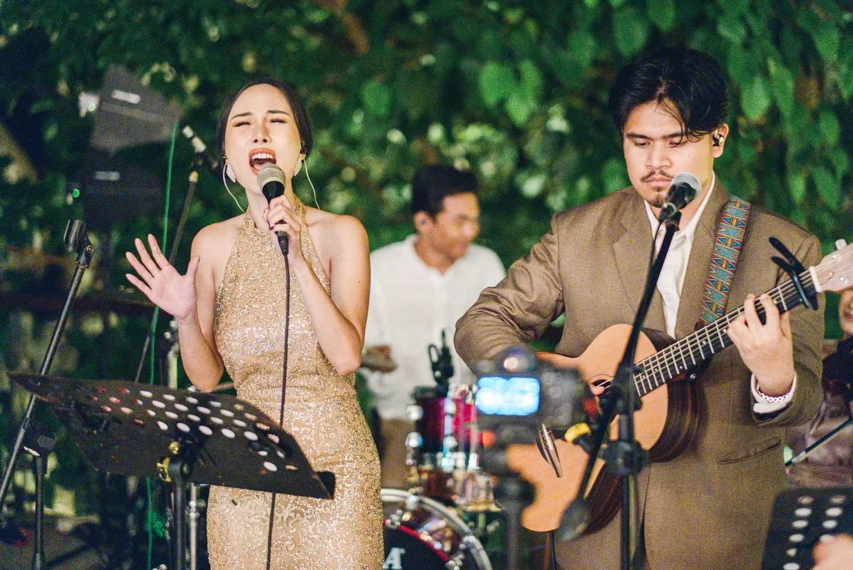La Oficio band performs live with a singer, guitarist, and drummer, set against a backdrop of lush greenery.