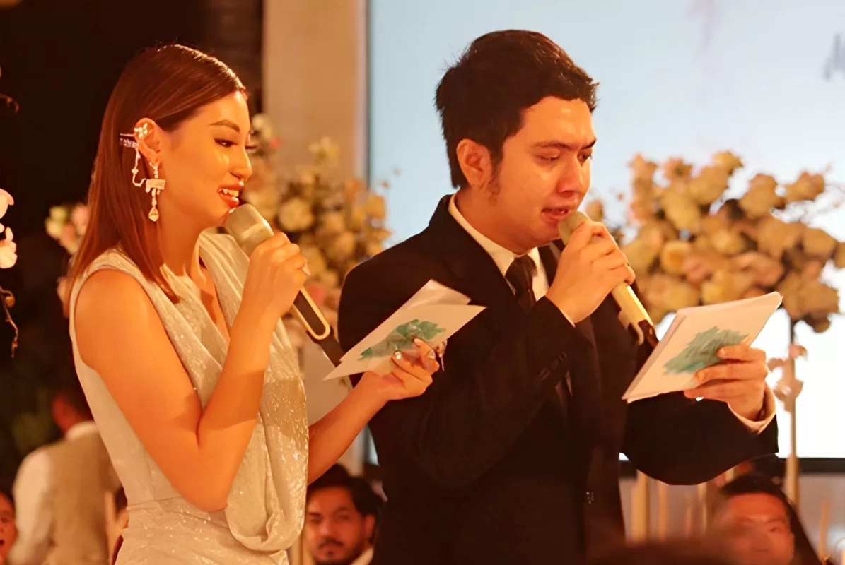 MC Bayu and partner MC speaking at a wedding reception in Tirtha Bali, holding microphones and reading notes on stage.