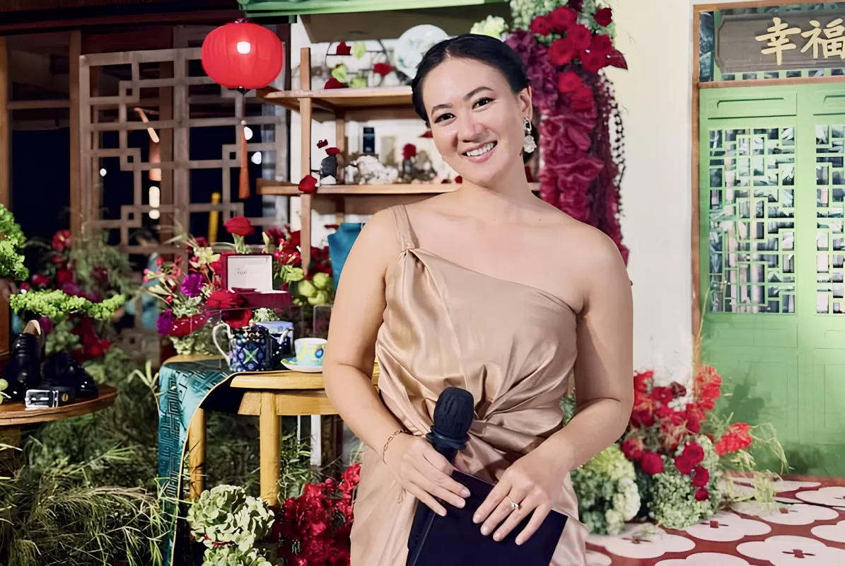 Tirza, a professional MC, is holding a microphone and cue card at a decorated indoor event setting.