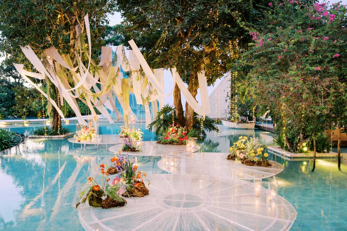 A dreamy eco-conscious wedding setup at Tirtha Bali featuring floating floral islands, flowing fabric drapes, and twinkling fairy lights suspended from trees, all surrounded by serene turquoise water and lush greenery.