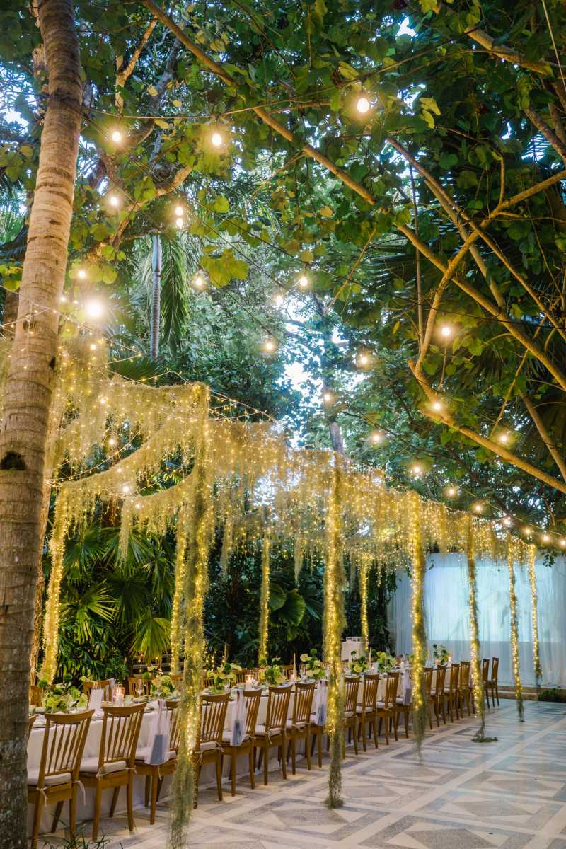 An elegant garden wedding reception setup at The Glass House, with long wooden tables under twinkling string lights and lush trees.