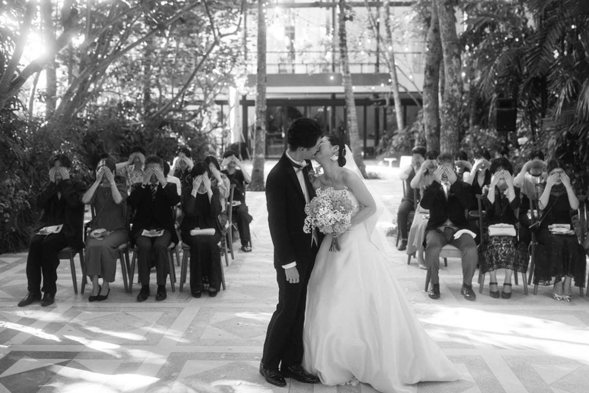 A fun wedding moment unfolds as the bride and groom kiss while guests hide their faces at Tirtha Bali.
