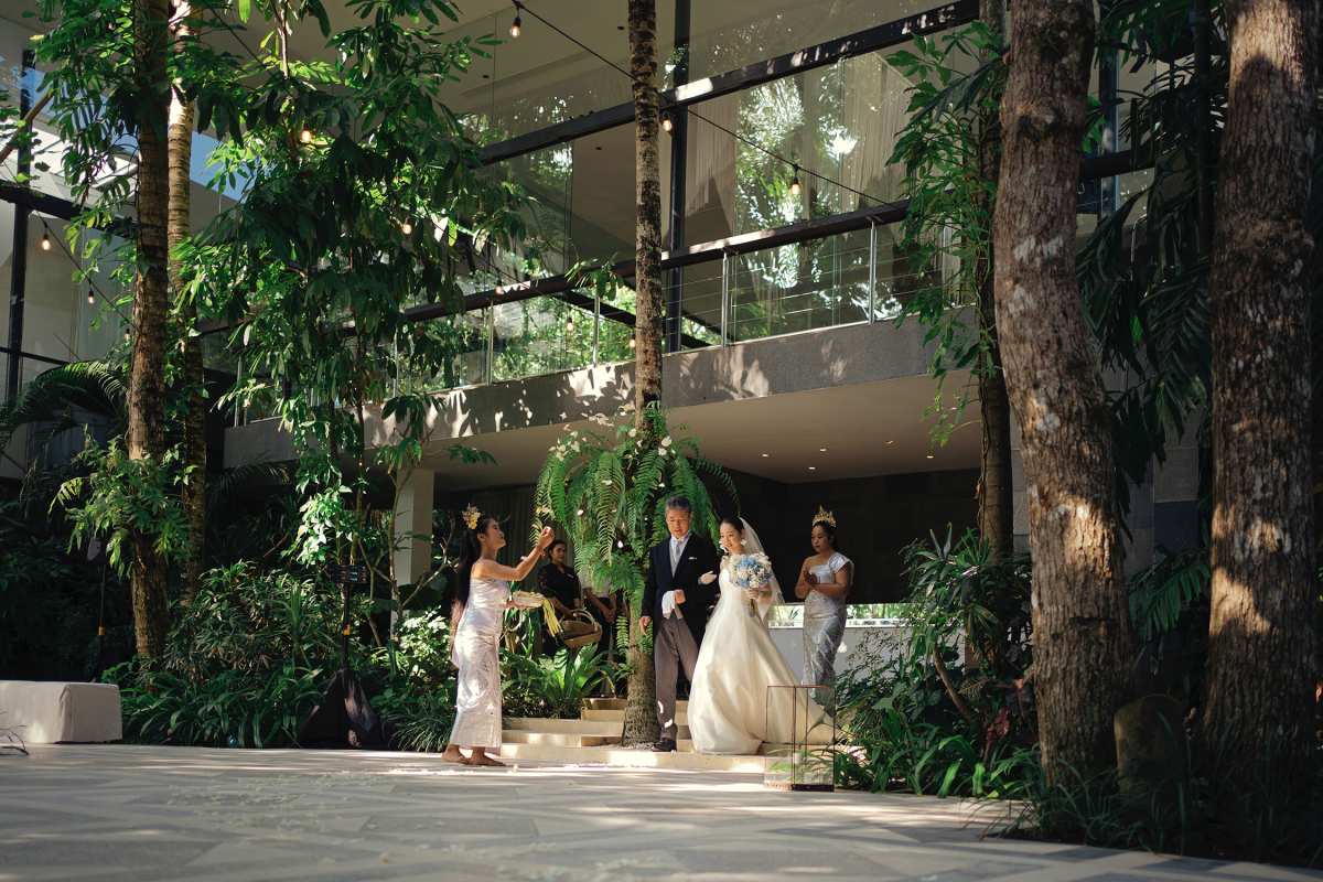 The bride walks down the aisle with her father under lush tropical greenery at Tirtha Bali.