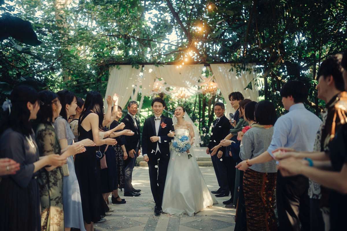 Bride and groom walk through cheering guests with falling petals at Tirtha Bali’s garden venue.