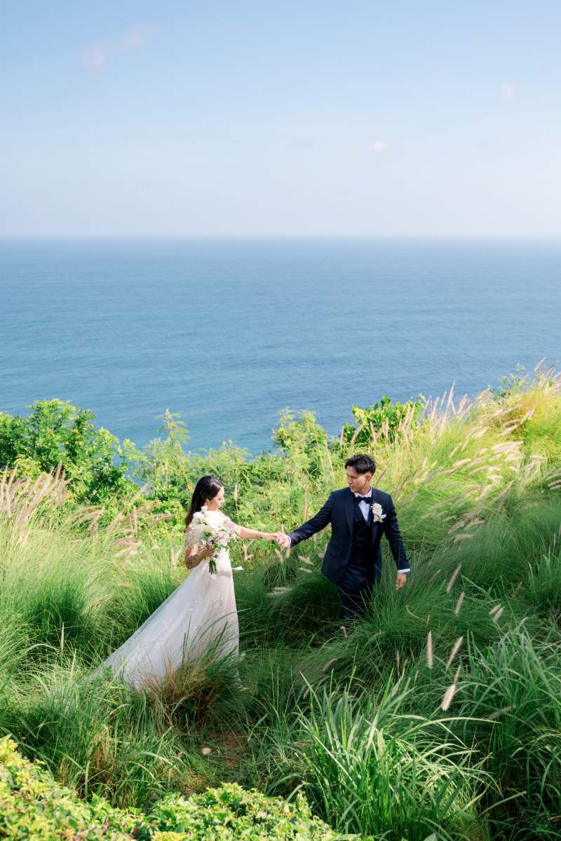Bride and groom walking through tall grass with ocean views at Tirtha Uluwatu.