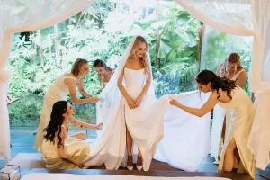 Bride in a white gown is helped by bridesmaids as they adjust her veil and train in the Glass House by Tirtha master bedroom.
