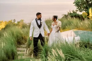 Bride holding a bouquet, walking hand in hand with the groom, with the ocean gently peeking through in the background.