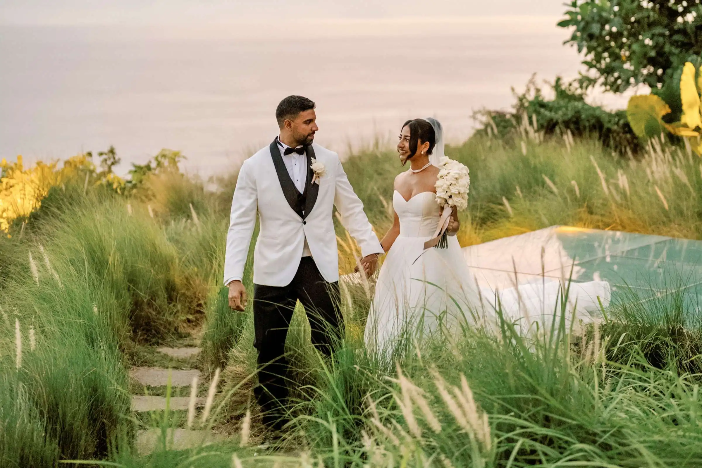 Bride holding a bouquet, walking hand in hand with the groom, with the ocean gently peeking through in the background.