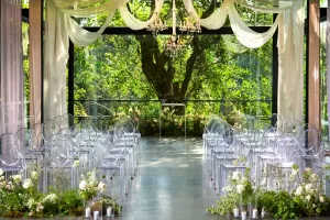 Wedding ceremony setup at Plumeria Room with greenery beyond glass walls, crystal chandeliers, and clear seating.