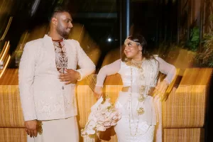 Tam and Lash are smiling at each other in Sri Lankan wedding attire, bathed in golden light, with Tam holding a bouquet.