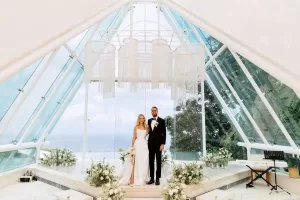 A couple at the altar of Glass Chapel by Tirtha Uluwatu, adorned with white floral arrangements under a fringe chandelier.