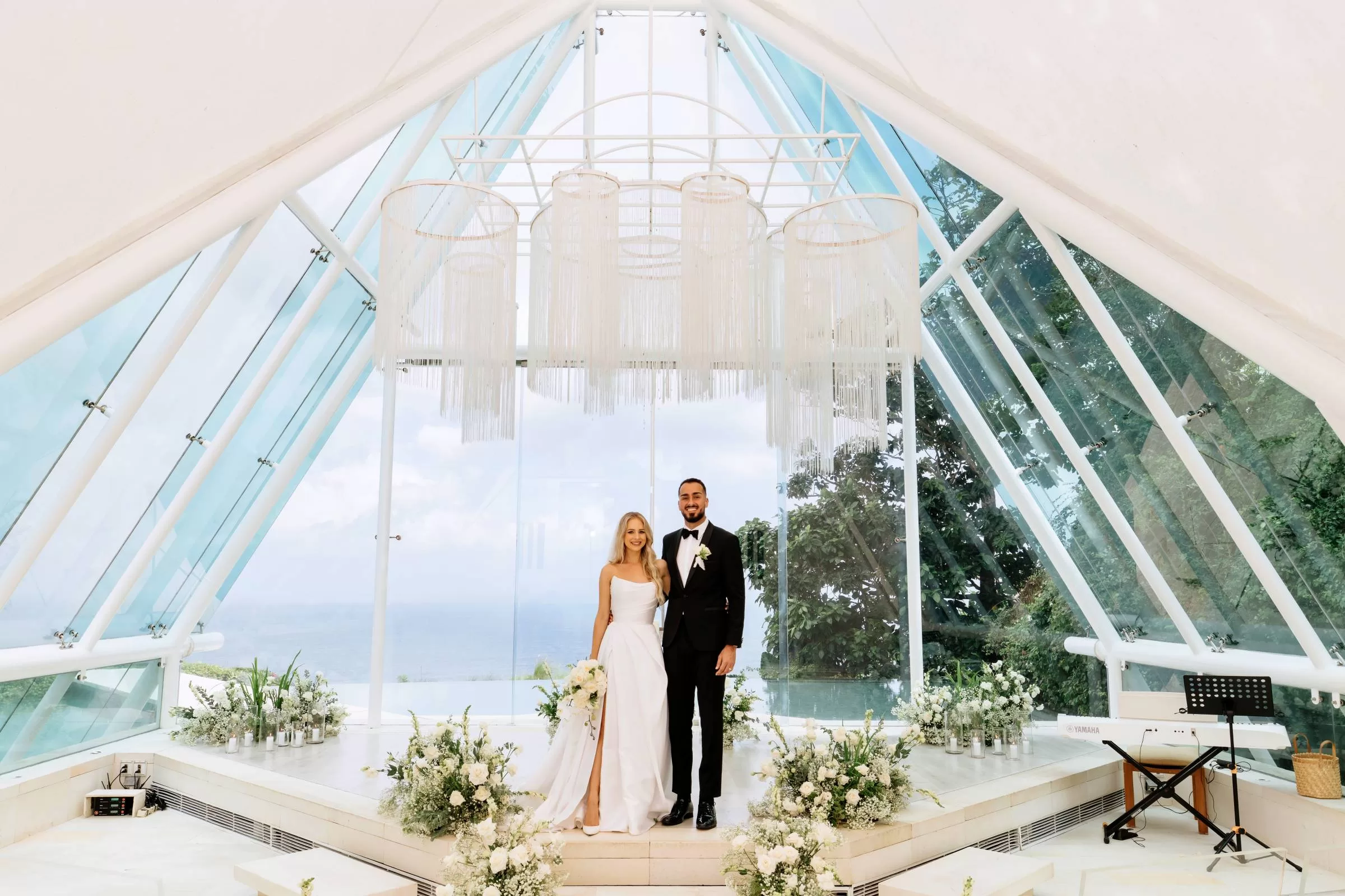 A couple at the altar of Glass Chapel by Tirtha Uluwatu, adorned with white floral arrangements under a fringe chandelier.