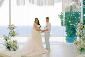 A couple holds hands and smiles at the guests, standing at a floral-decorated altar beneath a chandelier with an ocean view.