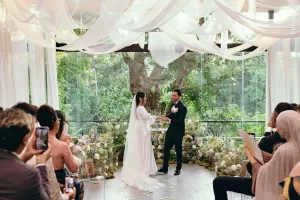 A couple holding hands at the Plumeria Room, surrounded by guests, florals, a draped ceiling, and a garden backdrop.