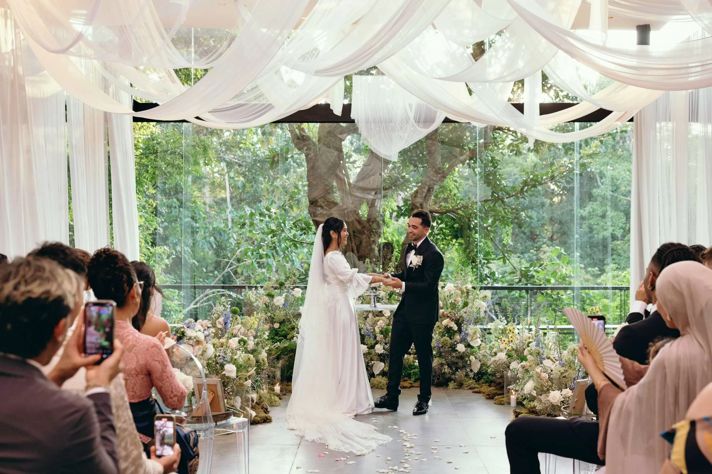 A couple holding hands at the Plumeria Room, surrounded by guests, florals, a draped ceiling, and a garden backdrop.