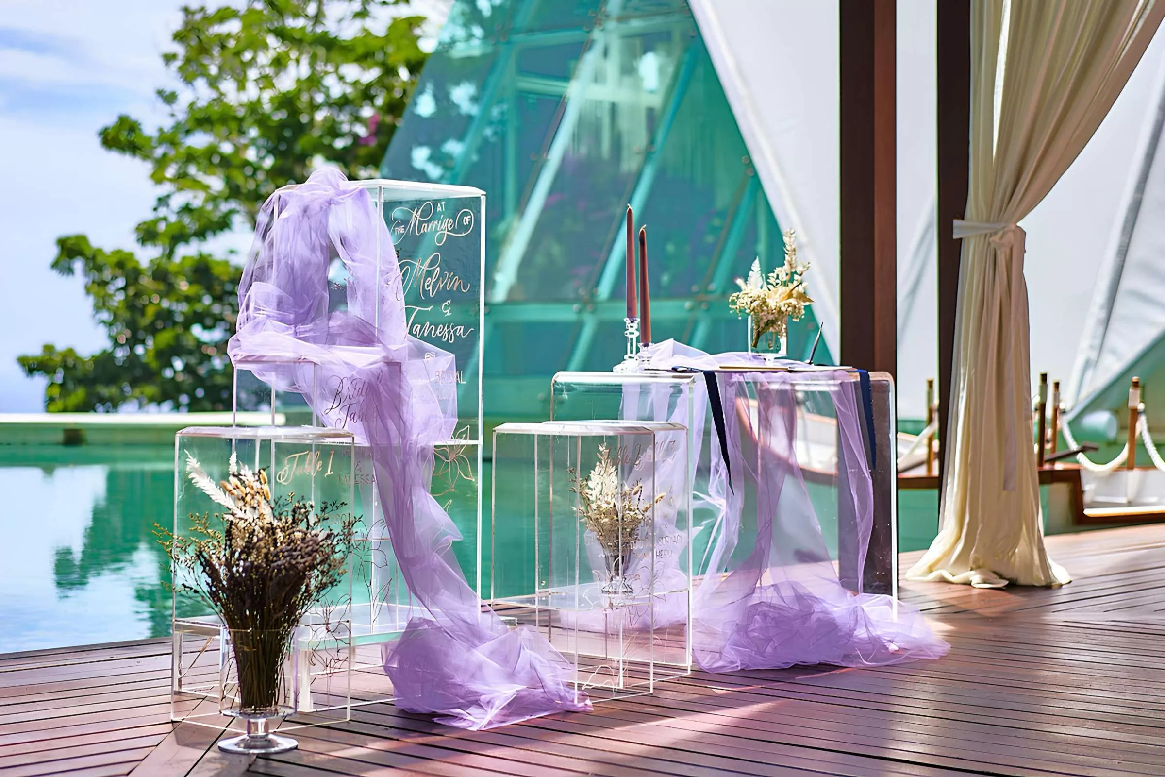 Acrylic welcome sign and pedestals by the poolside arranged with pastel purple tulle, flowers, and candles at Tirtha Uluwatu.