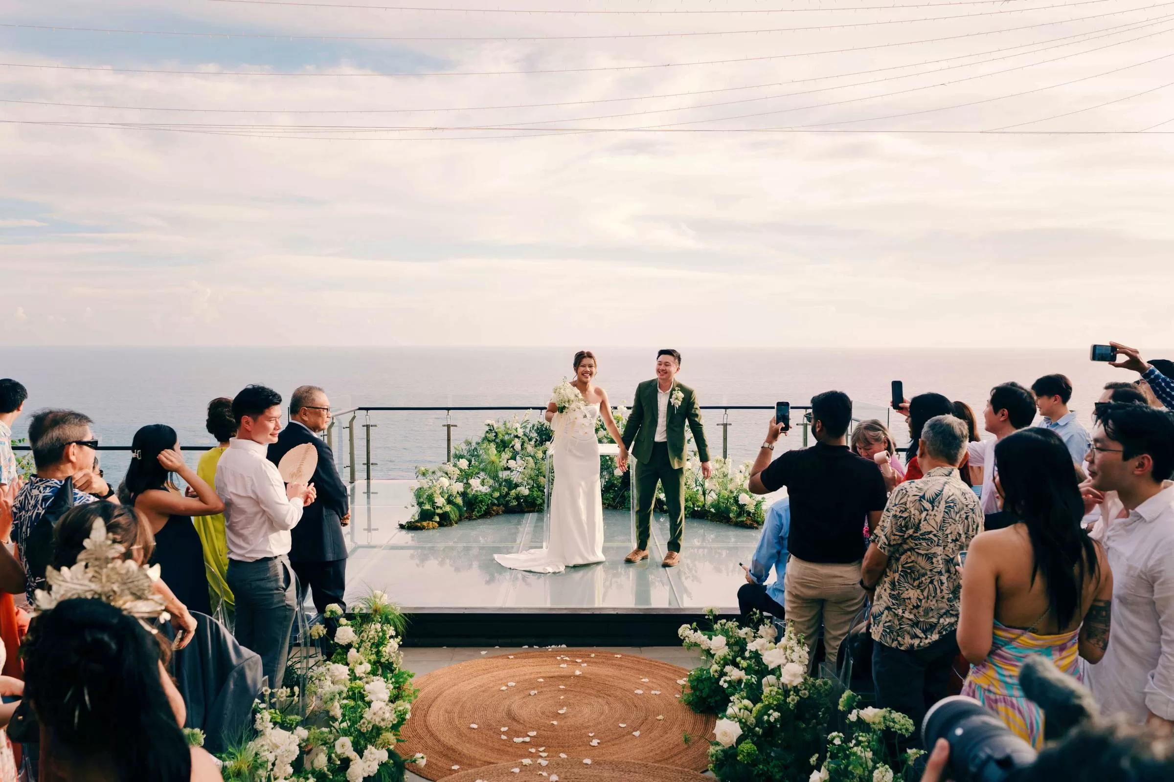 A newlywed couple hold hands on the Glass Stage at Tirtha Uluwatu with an ocean backdrop as the guests cheer and photograph.