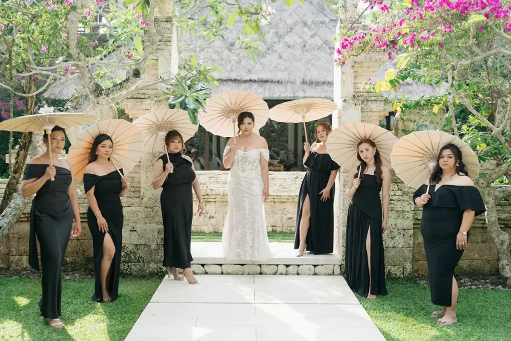 Eliza in her wedding dress with her bridesmaids in black dresses, all holding parasols at Tirtha Uluwatu's stone gate.