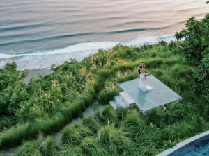 Couple slow dancing on a clifftop platform at Tirtha Uluwatu, surrounded by tall grass and overlooking the ocean.