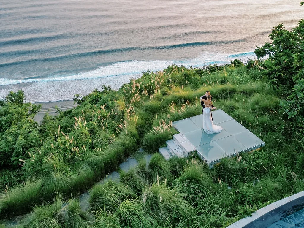 Couple slow dancing on a clifftop platform at Tirtha Uluwatu, surrounded by tall grass and overlooking the ocean.